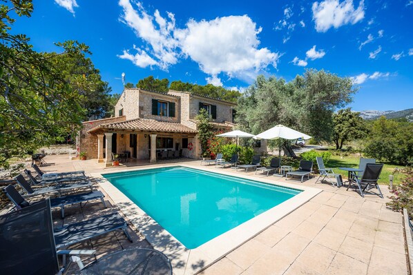 A pool with lounge chairs, a stone house with a tiled roof, and a clear blue sky.