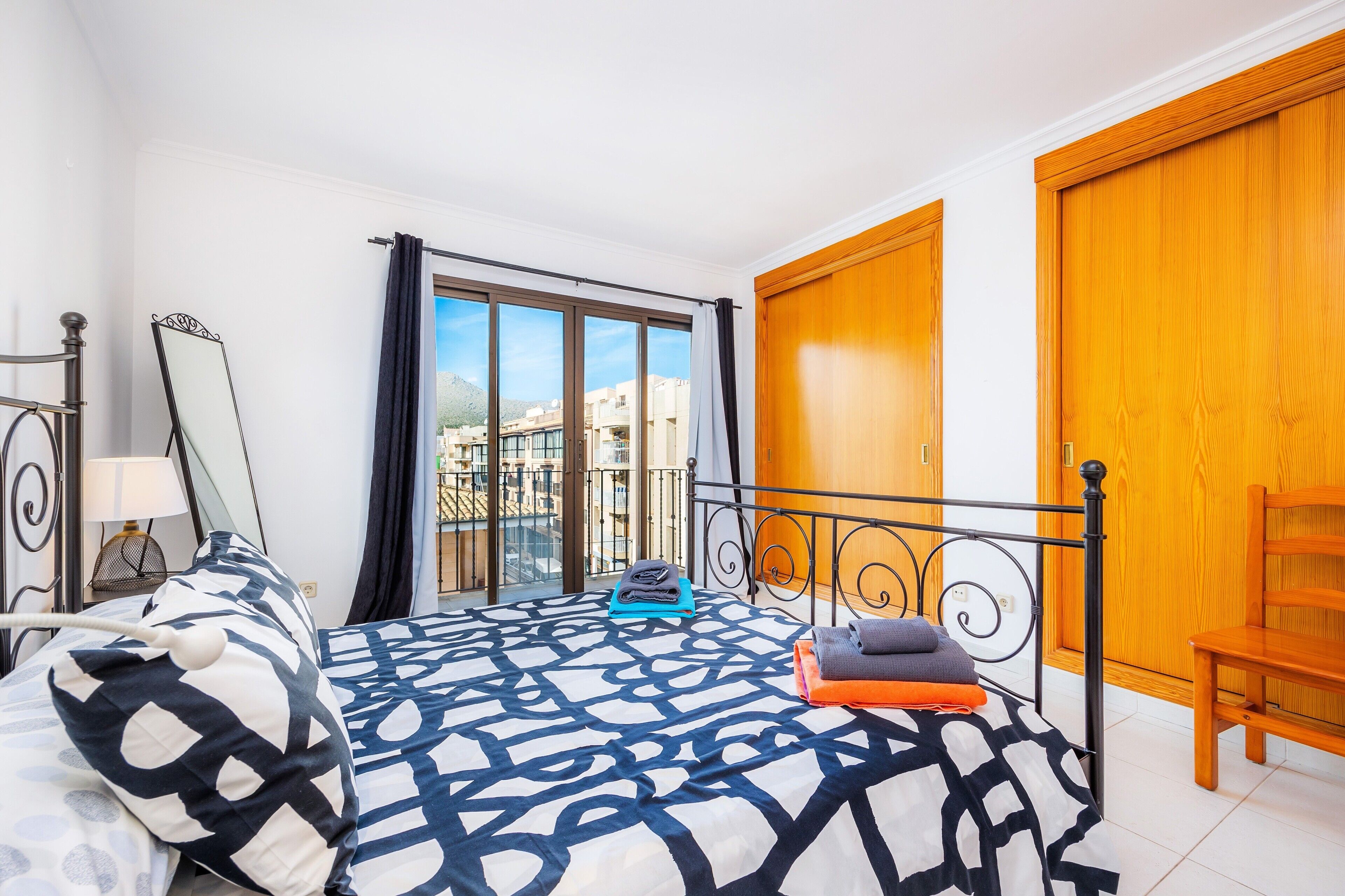 A bedroom with a bed, a nightstand, a mirror, a window with curtains, and a wooden door.