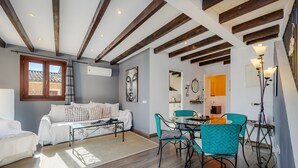 Una sala moderna con sofá, una mesita de centro y sillas color turquesa. Hay un comedor con mesa y sillas, y una cocina al fondo.