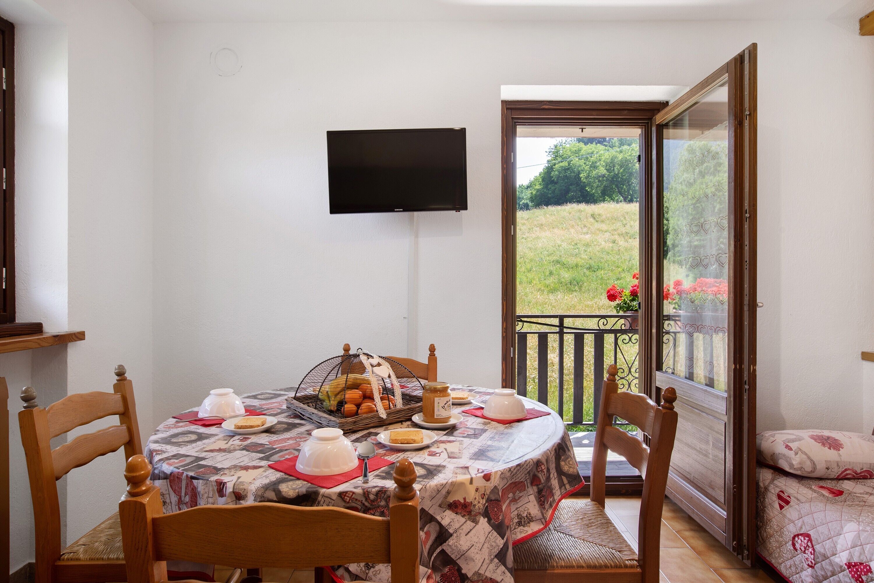 Zona pranzo con tavolo apparecchiato per due, televisore a parete e balcone con vista sul verde.