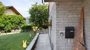A stone building with a stone staircase leading to a door, a wooden sculpture on the wall, and a well-maintained lawn with a tree and a bush. - Holiday Home 'Le Cré Appartamento 2' with Mountain View, Shared Garden and Wi-Fi (Introd)
