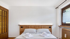 A hotel room with a wooden headboard, a bed with white linens, a grey blanket, and a window with a curtain. - Holiday Home 'Le Cré Appartamento 5' with Mountain View, Shared Garden and Wi-Fi (Introd AO)
