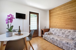A room with a wooden wall, a sofa, a TV, a dining table with chairs, and a potted plant.