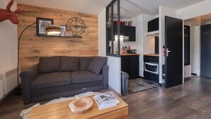 A modern living room with a grey sofa, wooden wall paneling, a mounted clock, and a kitchen area with a microwave and cabinets. - Studio "2 Alpes" with Mountain View, Private Terrace, and Balcony (Les Deux Alpes)