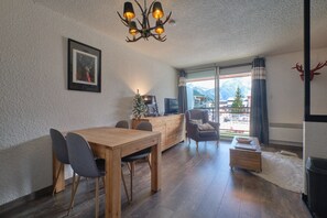 Un espace repas avec une table et des chaises en bois, un lustre, un téléviseur et une vue sur les montagnes grâce à de grandes fenêtres.