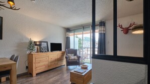 A modern living room with a wooden dining table, a sofa, a TV on a wooden cabinet, and a large window with curtains. - Studio "2 Alpes" with Mountain View, Private Terrace, and Balcony (Les Deux Alpes)