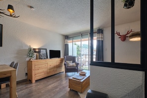 Un salon moderne avec une table à manger en bois, un canapé, un téléviseur posé sur un meuble en bois et une grande fenêtre avec des rideaux.