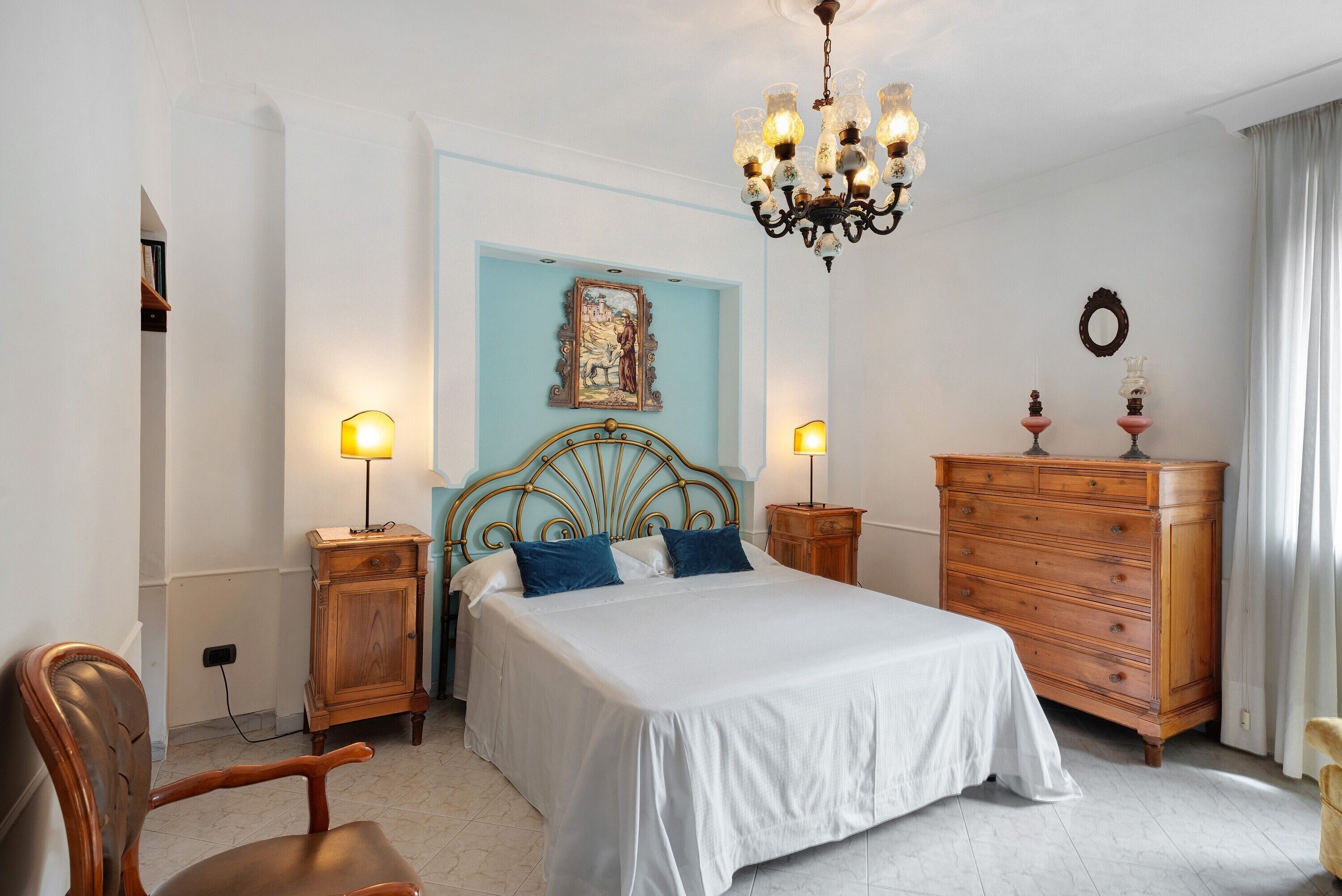 A bedroom with a bed, wooden dresser, nightstands, a chair, a chandelier, and a painting on the wall.