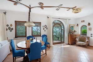 A dining area with a wooden table, blue chairs, a chandelier, and a view through large windows. - Villa 'Simba' with Sea View, Wi-Fi and Air Conditioning (Massa Lubrense)