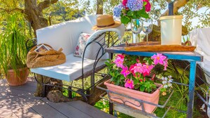 Un espacio de estar al aire libre con muebles blancos, un bolso tejido, un sombrero y una mesa con un jarrón de flores y un vaso.