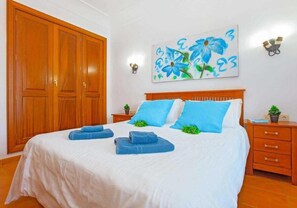 A bedroom with a large wooden wardrobe, a bed with blue pillows and towels, a painting of flowers on the wall, and a nightstand with a plant. - 2 Bedroom, 2 Bathroom Villa in Great Location (Playa Blanca)
