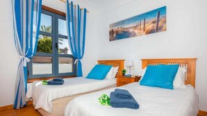 A hotel room with two beds, blue curtains, a painting of a beach, and a window with a view. - 2 Bedroom, 2 Bathroom Villa in Great Location (Playa Blanca)