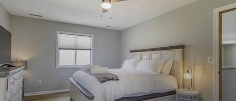 Un dormitorio con cama, ventilador de techo, mesita de noche con lámpara y ventana.