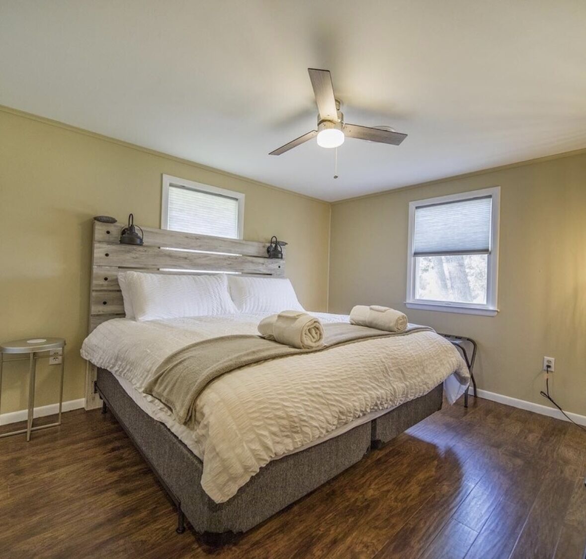 A bedroom with a wooden headboard, a bed with white linens, a ceiling fan, and two windows.