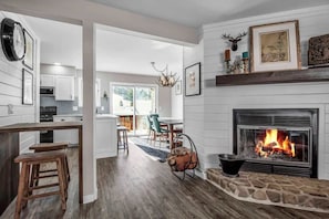 A modern kitchen with white cabinets, a dining area with a chandelier, and a fireplace with a fire. - Modern Townhome, 2 Decks, Fireplace (Ellicottville)