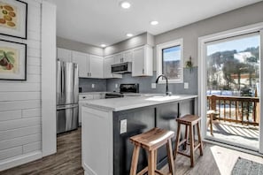 A modern kitchen with white cabinets, stainless steel appliances, a large island with stools, and a view of a snowy landscape through a glass door. - Modern Townhome, 2 Decks, Fireplace (Ellicottville)