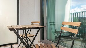 A balcony with a wooden table and chair, a glass door, and a view of the sky and buildings. - Cozy apartment in Oostende with WiFi (Oostende)