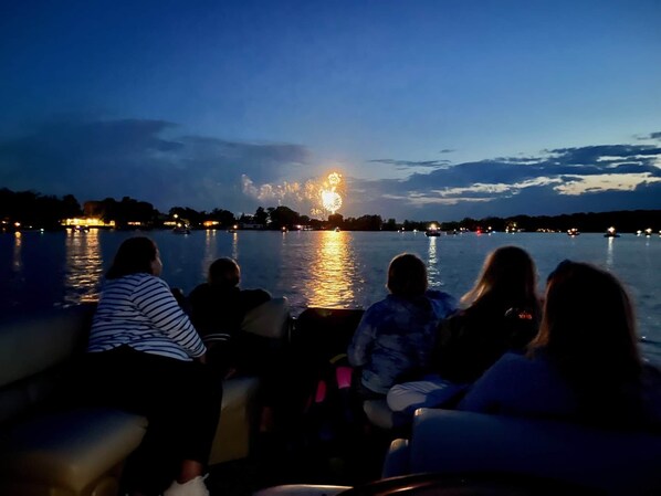 A group of people seated on a boat, facing a body of water with a fireworks display in the distance.