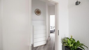 Ein Flur mit weißer Wand, Holzboden, einem weißen Schminktisch, einer Uhr, einem Topfpflanzen und einem Bett im Hintergrund.