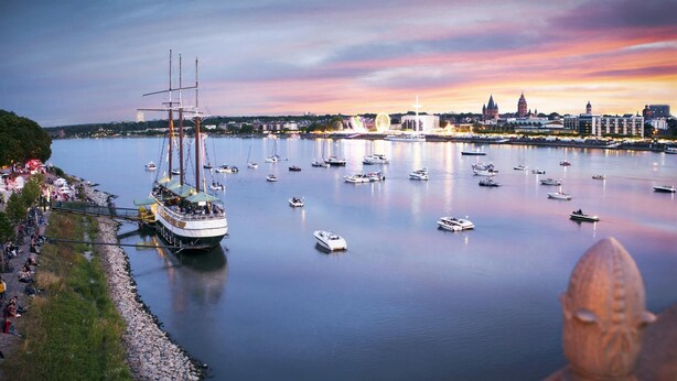 Ein Hafen mit mehreren Booten, einem großen Segelschiff und einer Stadtlandschaft im Hintergrund.