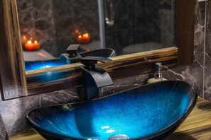 A bathroom with a blue glass sink, a wooden vanity, and a mirror reflecting a candle-lit background.