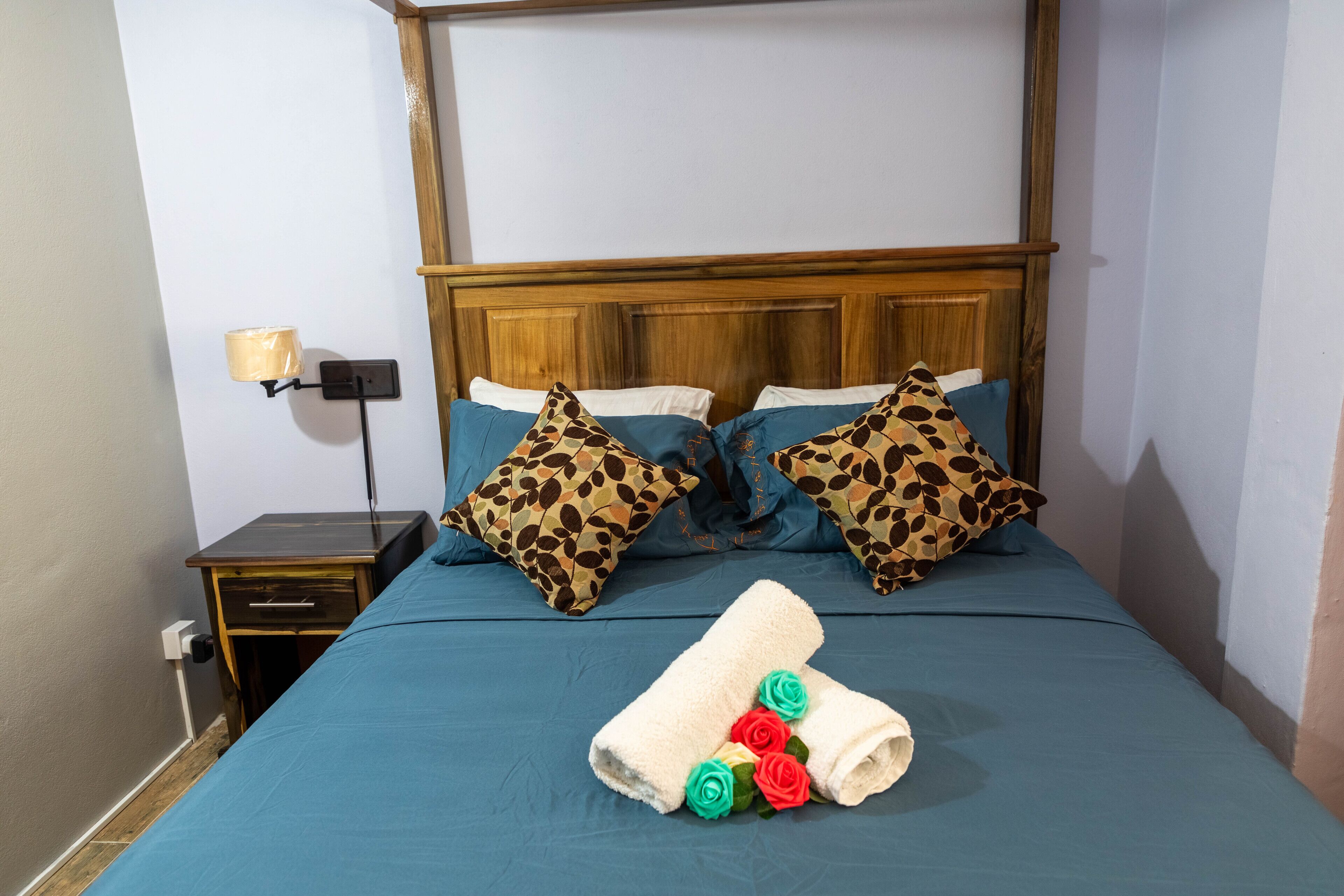 Een keurig opgemaakt bed met blauw beddengoed, twee kussens met luipaardprint, een houten hoofdbord en een nachtkastje met een lamp.