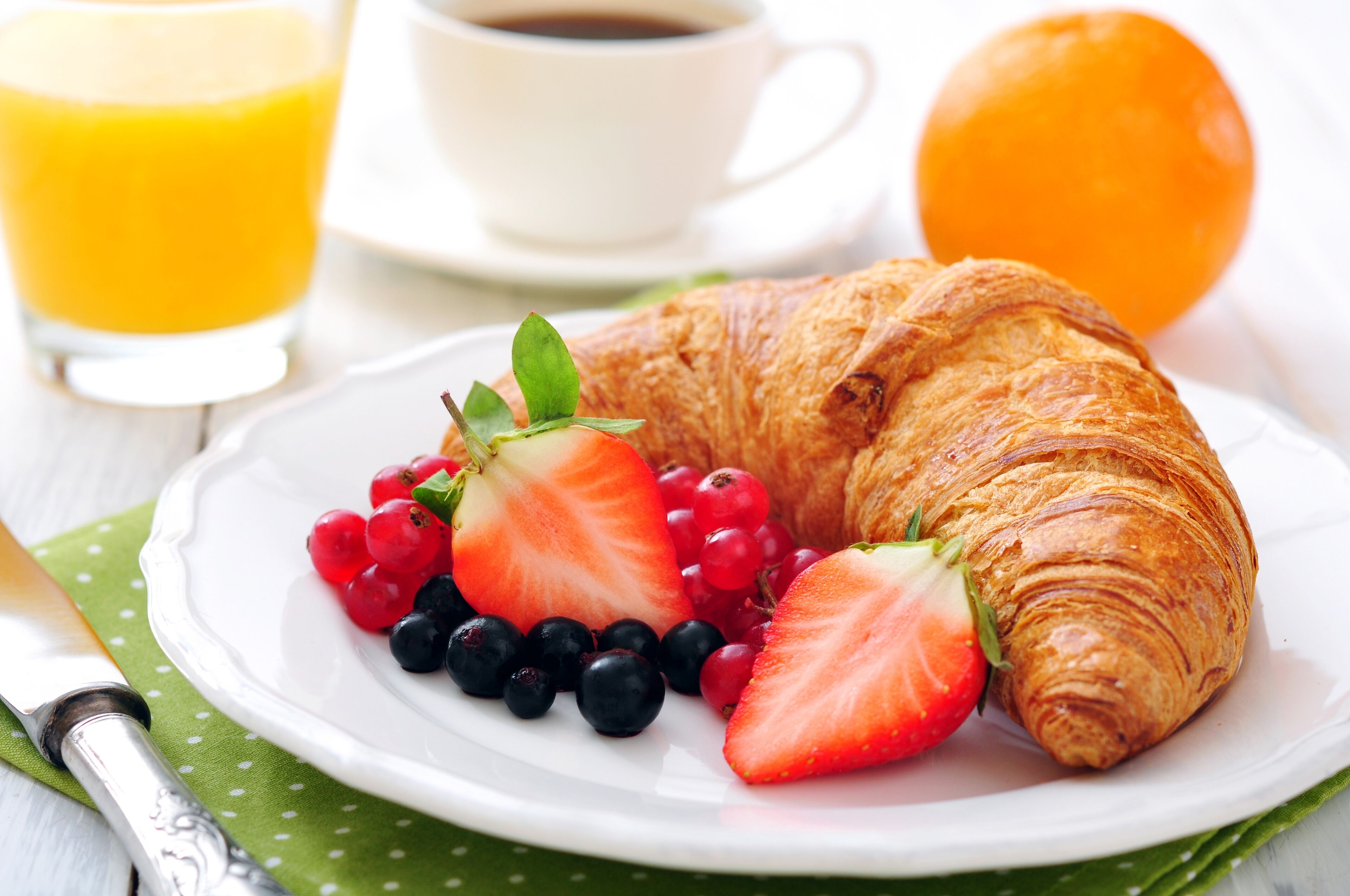Une assiette de petit-déjeuner comprenant un croissant, des fraises, des groseilles rouges et des myrtilles, un verre de jus d’orange et une tasse de café.