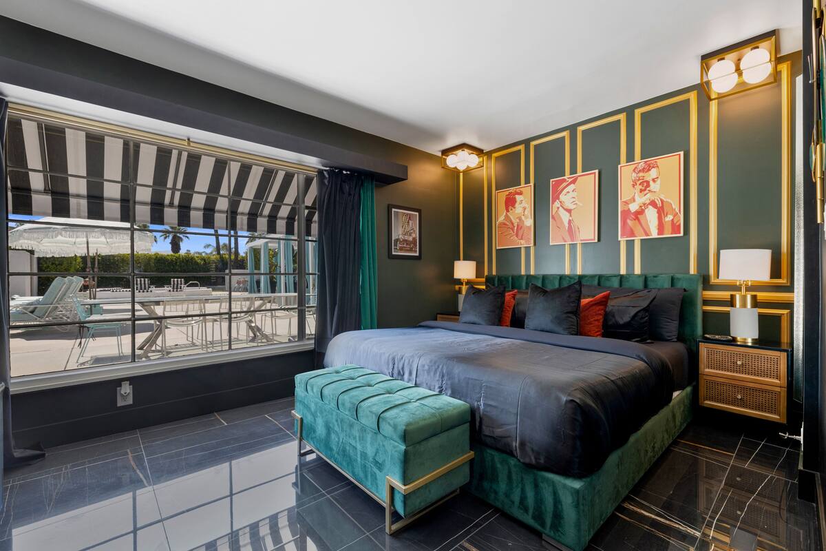 A modern bedroom with a large bed, a green upholstered bench, a view of an outdoor pool area, and a nightstand with a lamp.