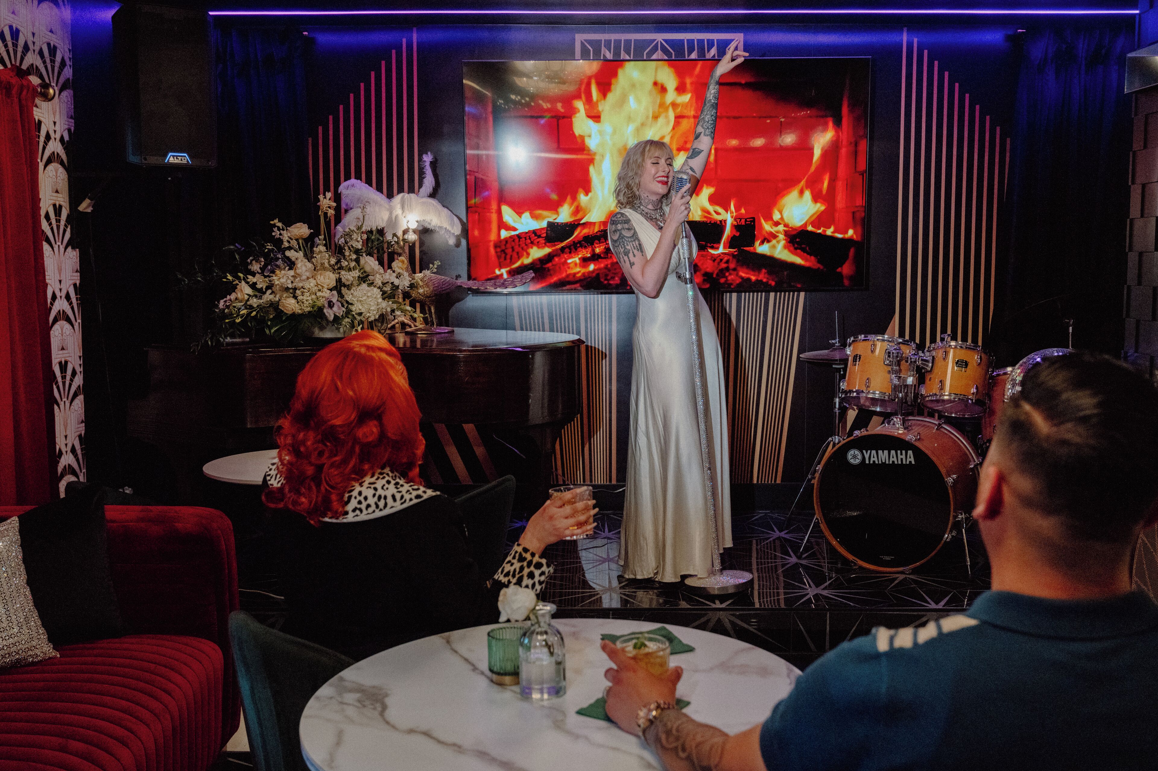 A person standing in front of a drum set with a Yamaha logo, a fire effect on the wall, and a table with drinks and a vase of flowers.