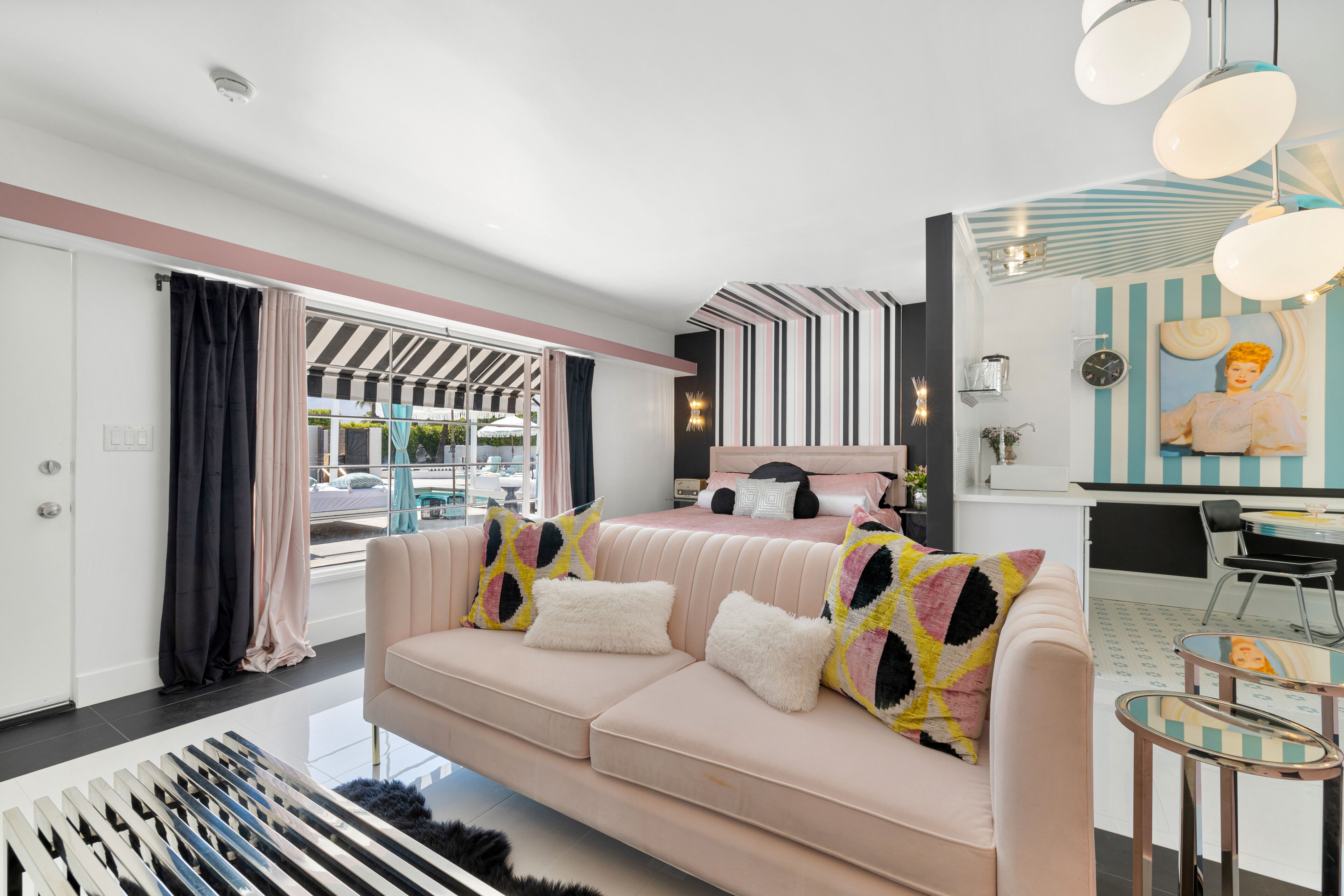 A modern hotel room with a large window, a sofa with patterned cushions, a bed with a striped headboard, and a small dining area with a chair and table.