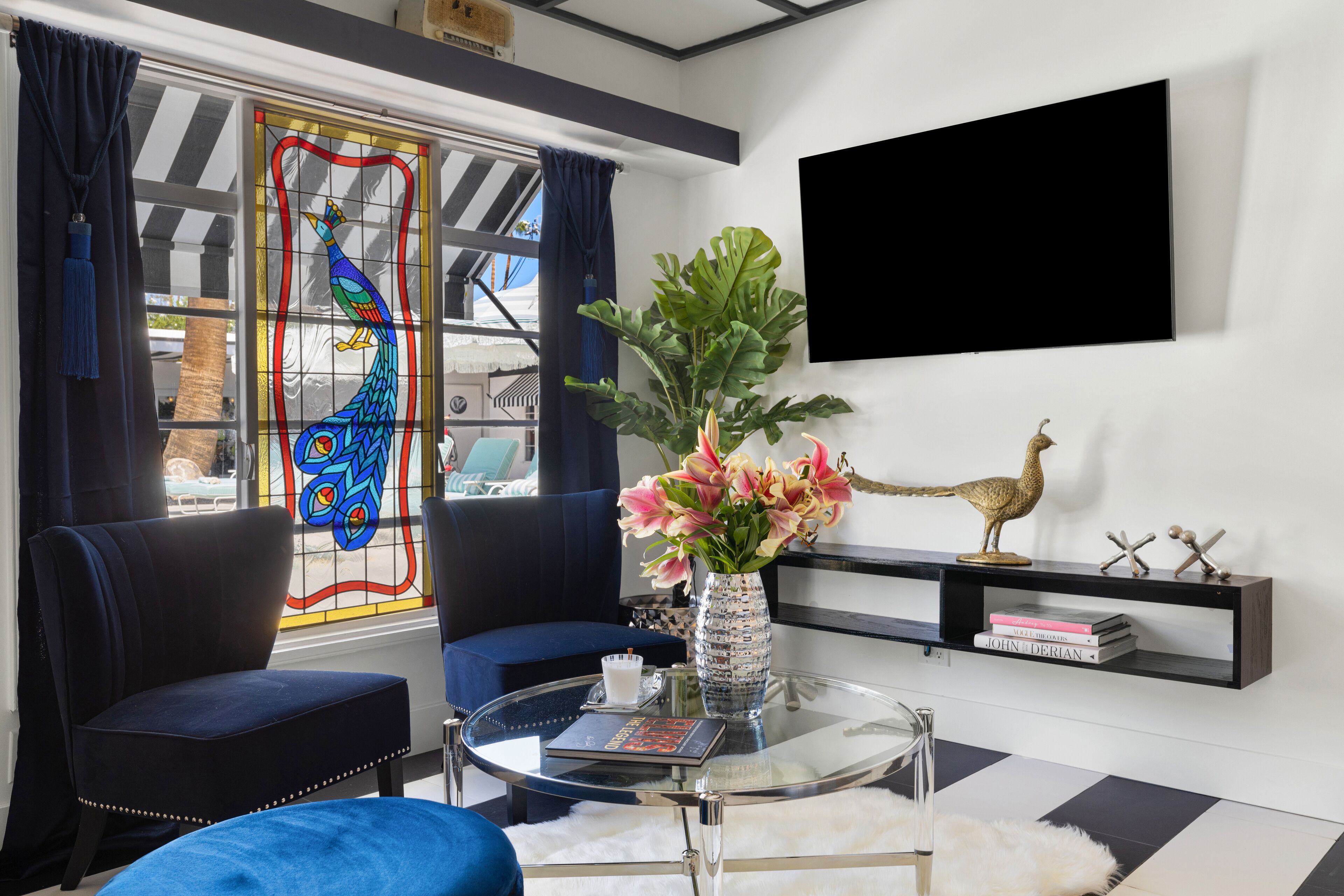 A modern living room with a glass coffee table, a blue sofa, a decorative vase with flowers, and a mounted TV.