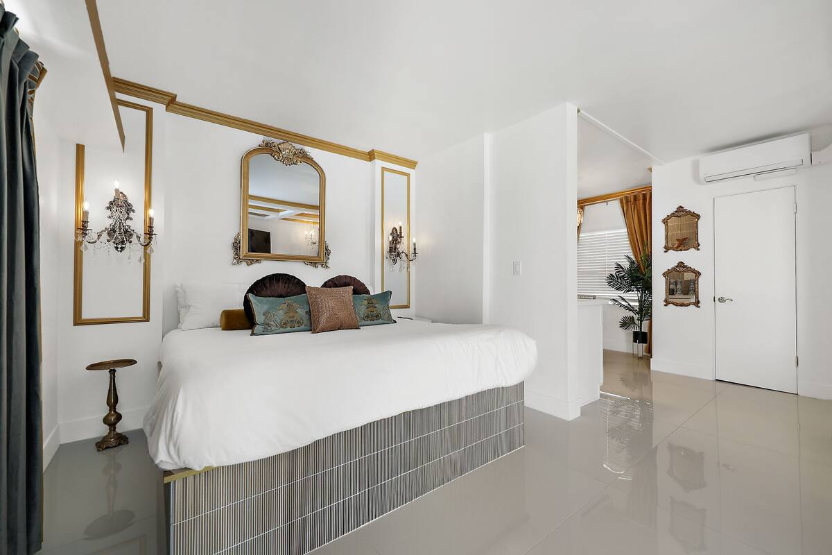 A bedroom with a large bed, a chandelier, a mirror, and a door leading to another room.