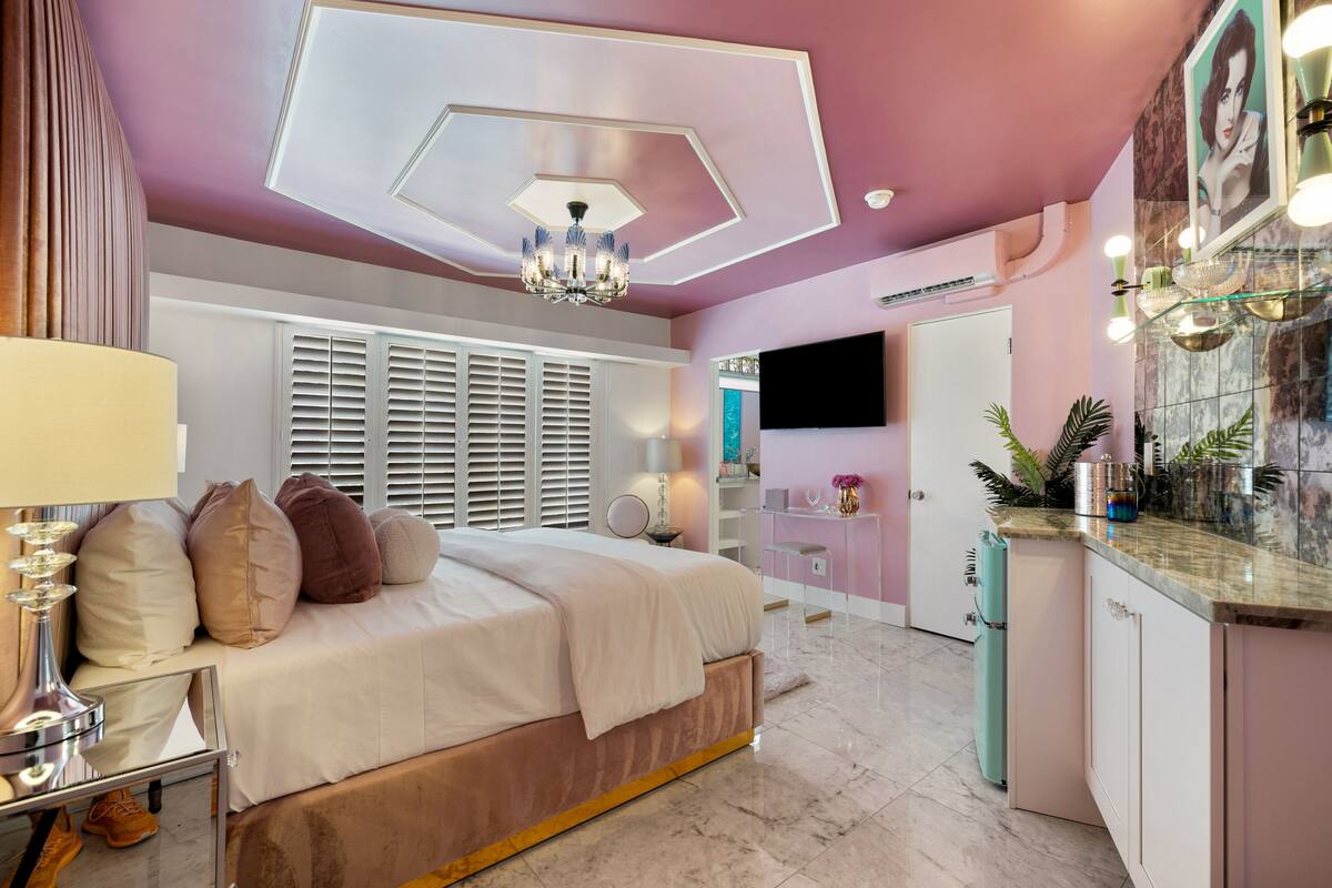 A bedroom with a bed, bedside table, and a mirror. There is a window with blinds, a ceiling light, and a TV mounted on the wall.