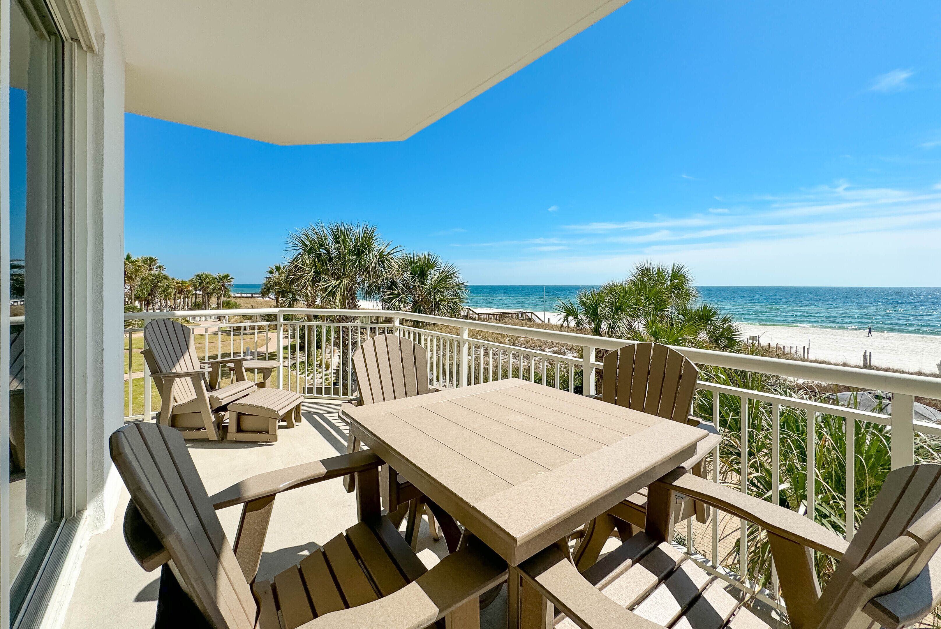 Ein Balkon mit Tisch und Stühlen, der Blick auf einen Strand mit Palmen und das Meer freigibt.