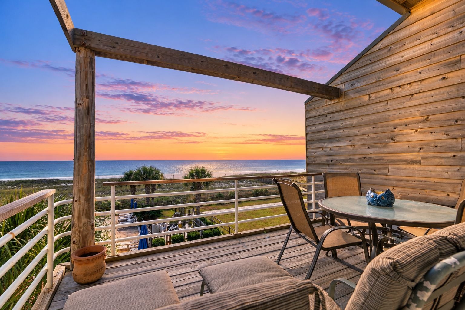 A wooden deck with a table and chairs, overlooking a beach at sunset.