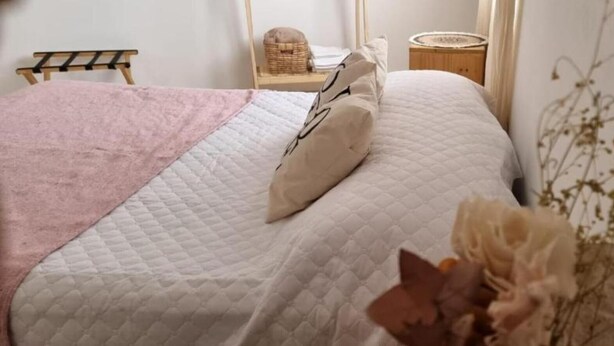 A neatly made bed with a white quilt and a pink blanket. There is a small bedside table with a basket and a round mirror.
