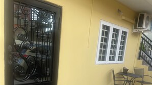 Eine gelbe Wand mit einem Metallgitter, ein Fenster mit weißen Rahmen und eine geflieste Terrasse mit Stühlen.