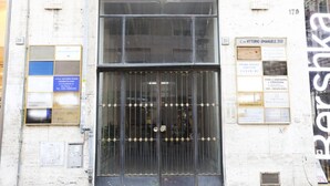 Fachada de edificio con un gran portón metálico, una ventana con diseño de cuadrícula y varias señales en las paredes.