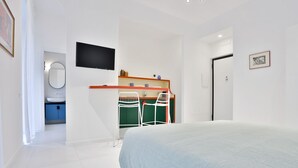 Un dormitorio con cama, televisor montado en la pared, escritorio con sillas y una puerta.