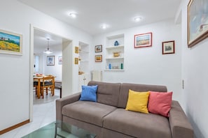 A living room with a sofa, glass coffee table, and wall-mounted shelves with framed pictures.