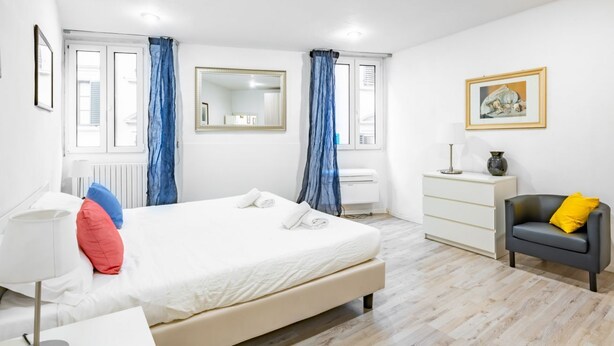 Un dormitorio con cama, cómoda, silla, lámpara, espejo y ventana con cortinas.