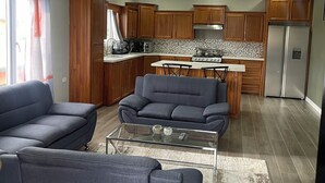 A modern living room with a grey sofa, a glass coffee table, and a kitchen in the background featuring wooden cabinets and a stainless steel refrigerator. - Charming 1-bedroom 1 bath apt. in awesome D'Abadie with AC, WiFi, Washer/Dryer. (D'Abadie)