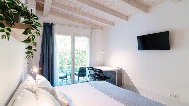 Une chambre avec un lit, un bureau, une chaise, une télévision et un balcon donnant sur un espace verdoyant.