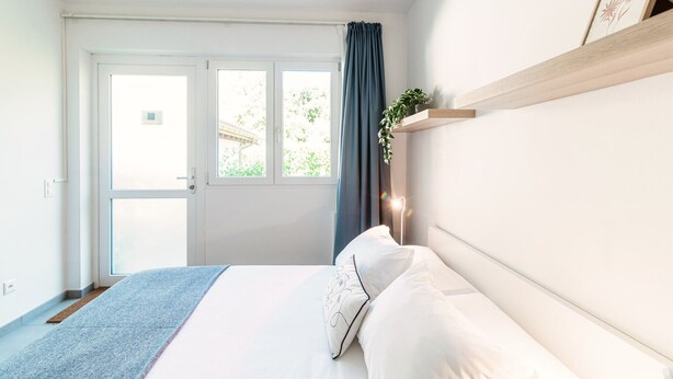 Een slaapkamer met een bed, een raam met gordijnen, een plant op een plank en een deur.