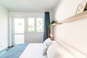 A bedroom with a bed, a window with curtains, a plant on a shelf, and a door. - Comfortable studio apartment near Origlio pond (Le Betulle Studio 2) (Origlio)