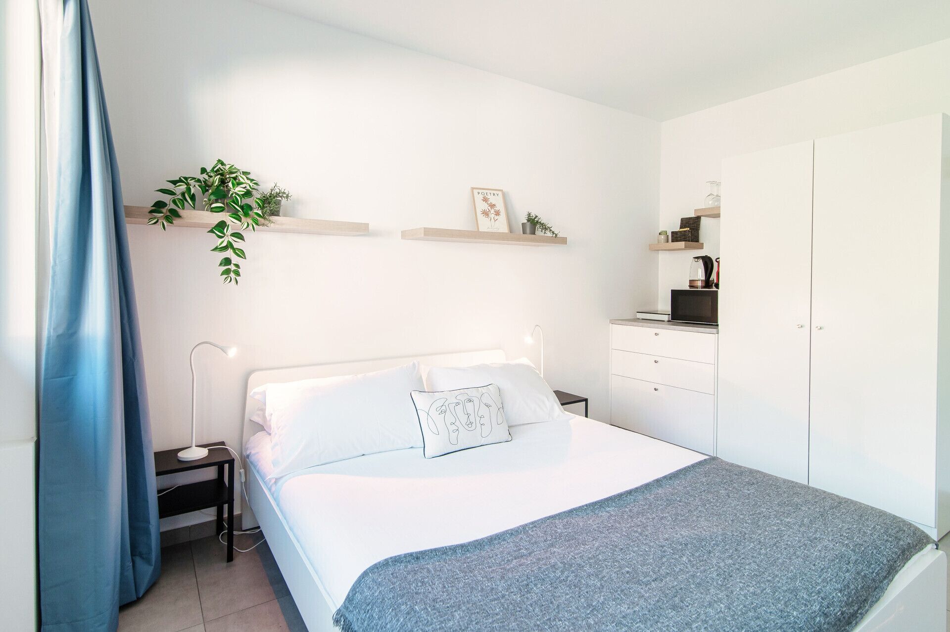 A bedroom with a bed, bedside table, lamp, and a small desk with a kettle.