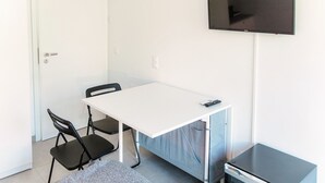 Een kleine kamer met een witte tafel, twee zwarte stoelen, een televisie aan de muur en een klein kastje.