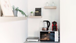 Een keukenhoek met een magnetron, waterkoker en koffiezetapparaat op een aanrecht. Daarboven staan twee houten planken met glazen en een kleine potplant.