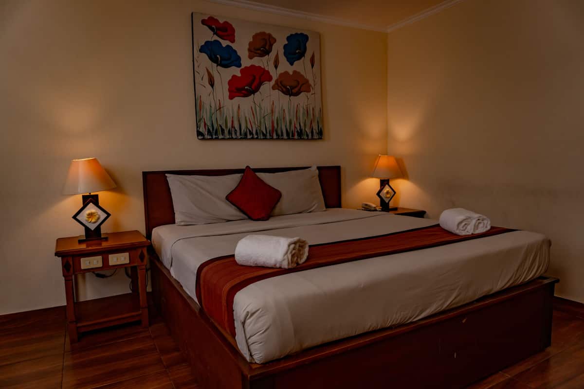A neatly made bed with a red pillow, two bedside lamps, and a floral painting on the wall.
