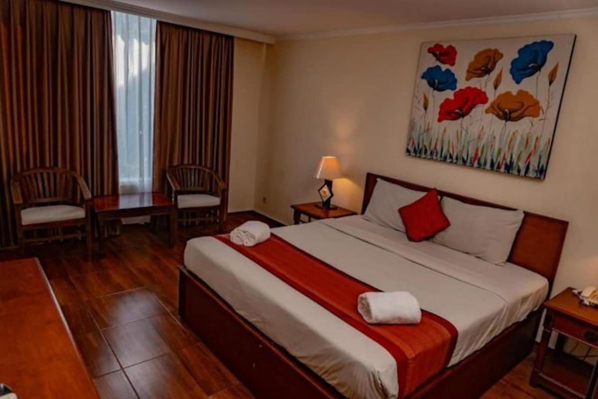A hotel room with a bed, a desk, two chairs, a painting, and a nightstand.